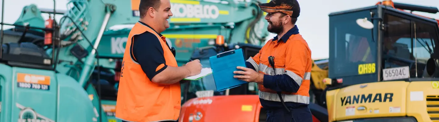 Two workers in uniform smiling while standing in front of construction equipment at a job site.