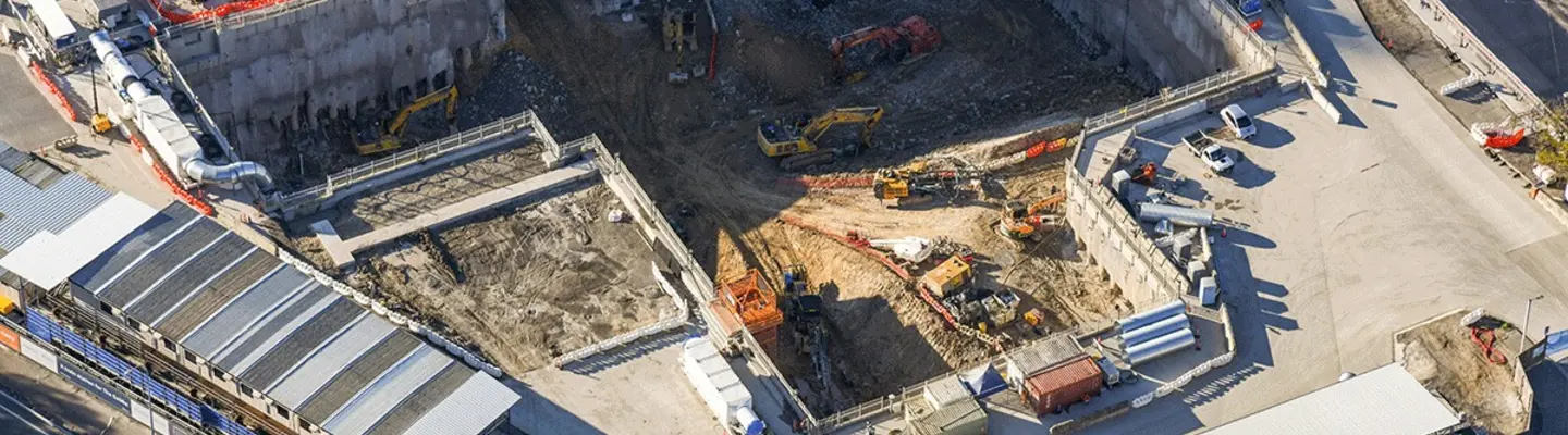 Various construction equipment working along a riverbank to build and reinforce a railway line with safety barriers.