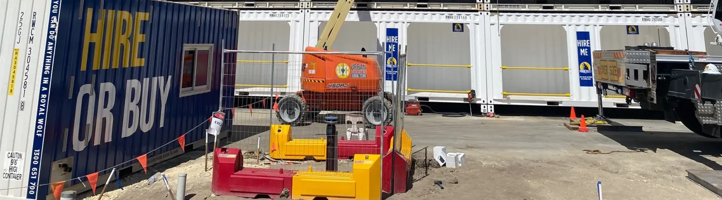 Construction site with shipping containers used as offices and storage, surrounded by safety barriers and workers in high-visibility gear.