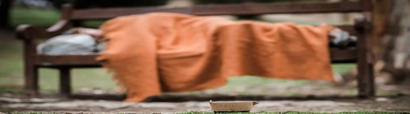 Homeless man sleeping on a bench in a park during the day, highlighting urban poverty and the need for shelter solutions.
