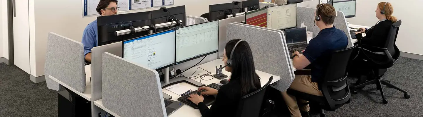Office staff working at computers in a modern workspace while a colleague updates a planning board on the wall.