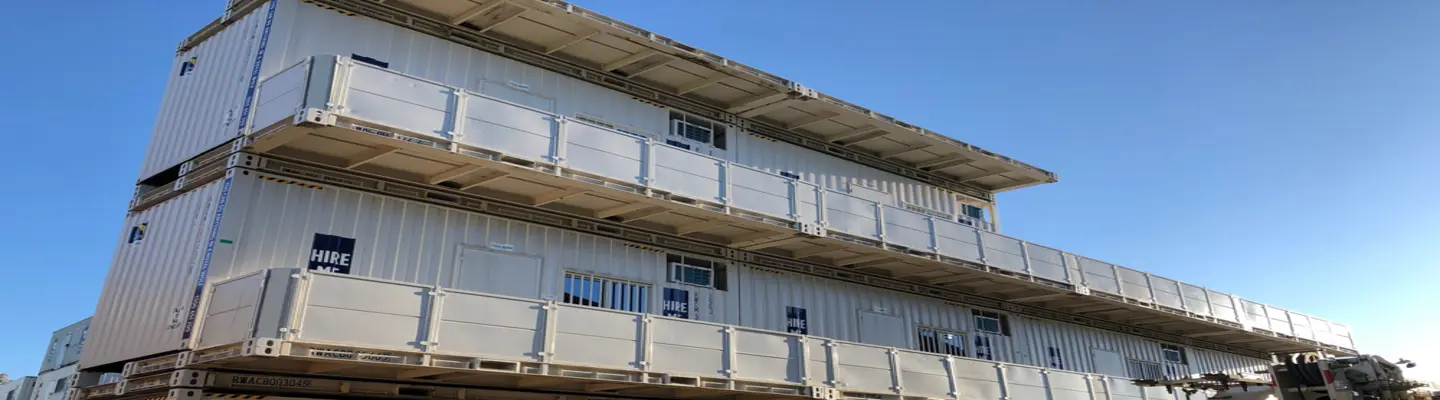 A multi-level structure made from stacked white shipping containers, featuring balconies and doors, set outdoors under a clear blue sky, with a digital timer display in the foreground.
