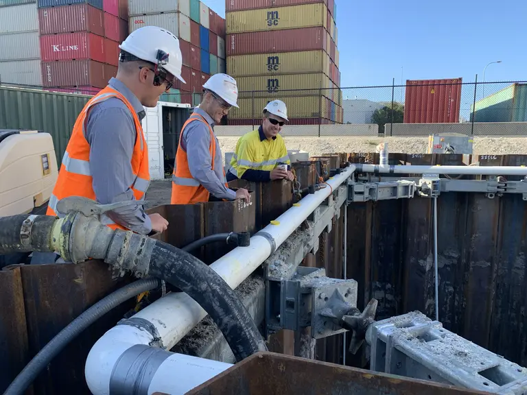 Three construction workers inspecting shoring and piping within a trench at an industrial site with shipping containers.