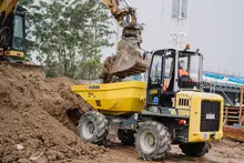 Man operating an excavator with attachment digging soil at a construction site with safety barriers in the background.