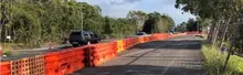 Orange construction barriers line a roadside, separating traffic from a closed lane with cars passing nearby.